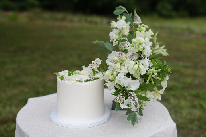 A cake decorated with a crown of white and off-white flowers beside a bouquet of the same.