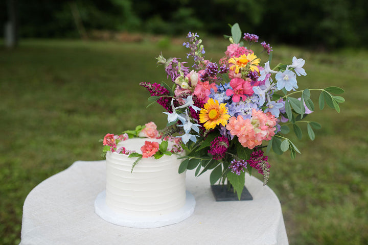 A cake decorated with a crown of bright, jewel-toned flowers beside a bouquet of the same.