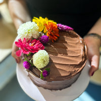 A person holding a round cake frosted with fudgey buttercream and decorated with a crescent of fresh flowers.
