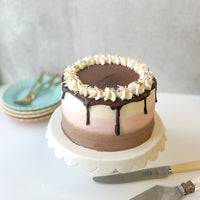 A round cake frosted with three bands of different flavors: vanilla, chocolate, and strawberry. The cake is topped with a chocolate ganache drip, a crown of whipped cream, and rainbow sprinkles.