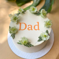 A cake frosted with smooth white buttercream and decorated with a crown of greenery. The word "dad" stenciled on top in orange.