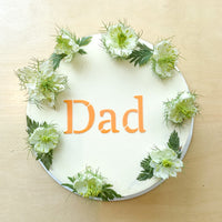 Top-down view of a cake frosted with smooth white buttercream and decorated with a crown of greenery. The word "dad" stenciled on top in orange.