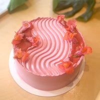 A cake frosted with hot pink buttercream. The sides of the cake are smooth, and the frosting is piped in a wavy pattern across the top of the cake; beside the waves are pools of rhubarb compote and fresh snapdragon flowers.