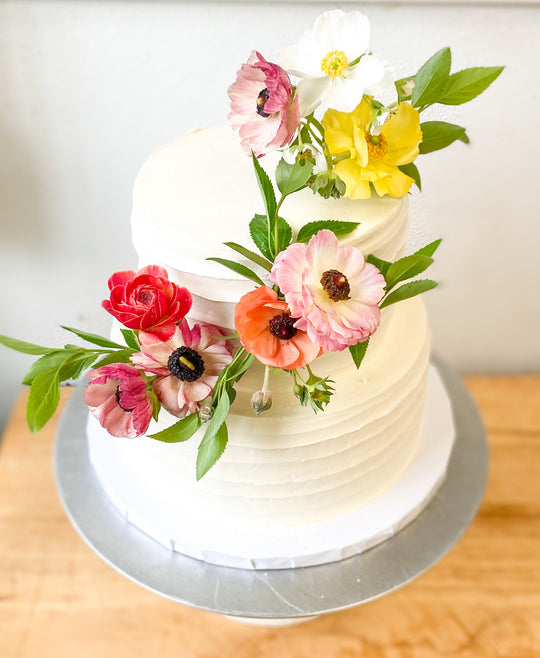 A two-tier cake decorated with a cascade of brightly colored fresh flowers. Colors include red, pink, and orange.