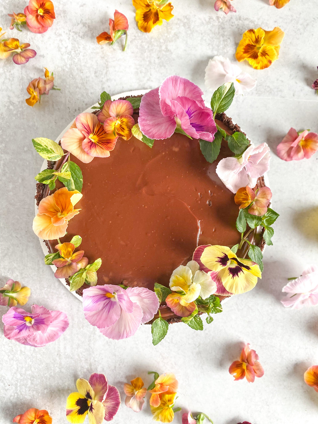 Top-down view of a cake covered in ganache and chocolate crumbs. The top of the cake is decorated with a crown of fresh flowers and greenery.