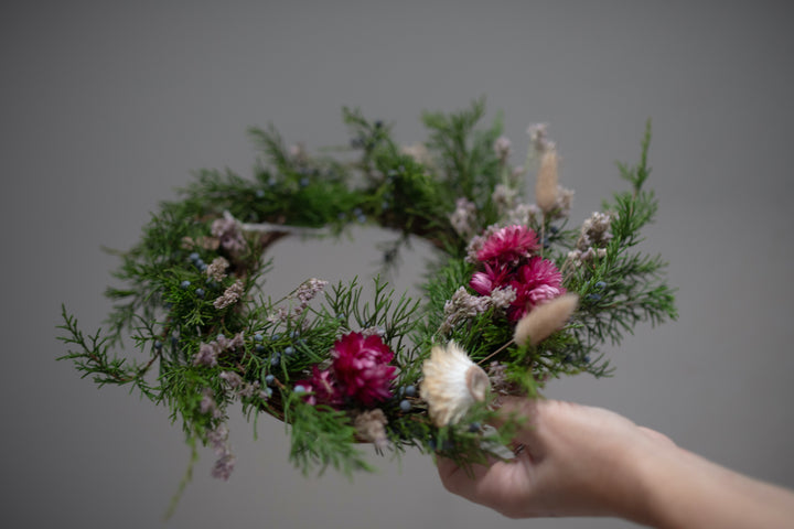 A woman holding a wreath made of evergreen fronds and dried flowers.
