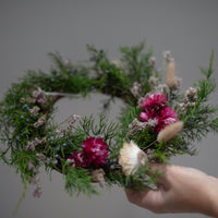 A woman holding a wreath made of evergreen fronds and dried flowers.