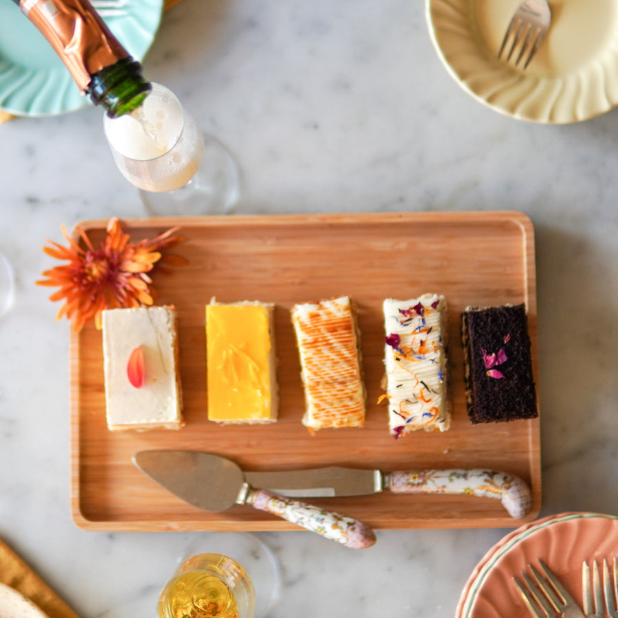 Five rectangular slices of cake, each a different flavor, arranged on a wooden platter alongside a knife and pastry server.