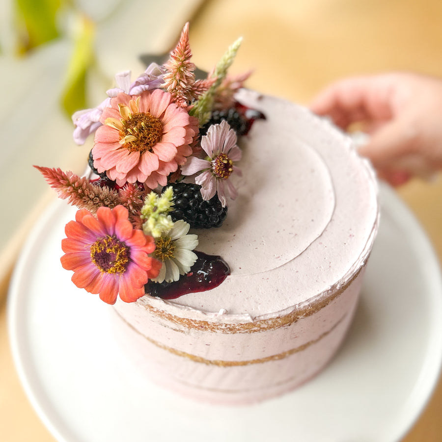 A round cake frosted with a sheer layer of purple buttercream and topped with a bouquet of flowers and blackberries.