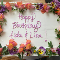 A white sheet cake with a crown of fresh flowers; "happy birthday Aida & Lina!" is piped in pink buttercream.
