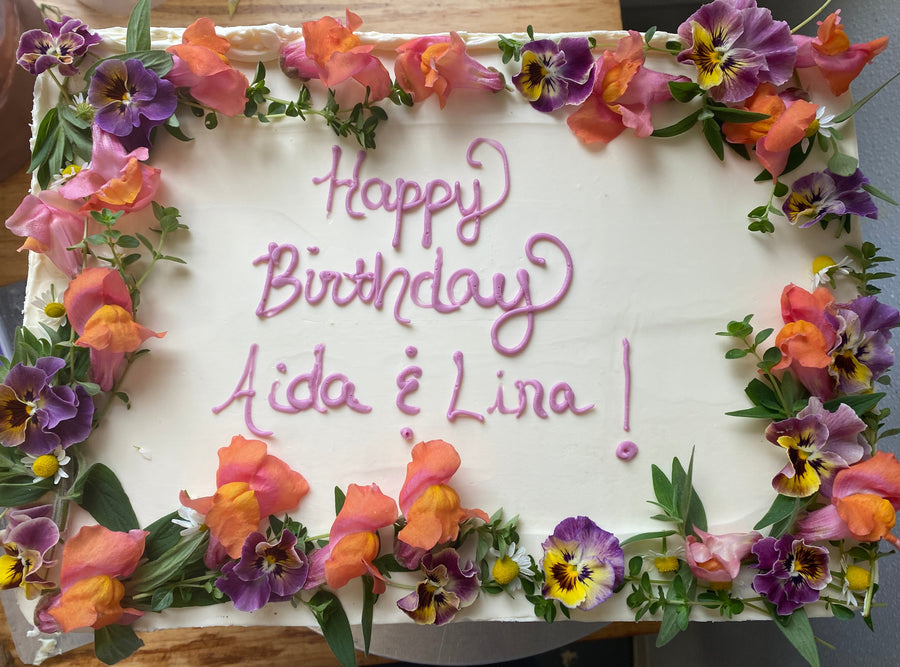 A white sheet cake with a crown of fresh flowers; "happy birthday Aida & Lina!" is piped in pink buttercream.