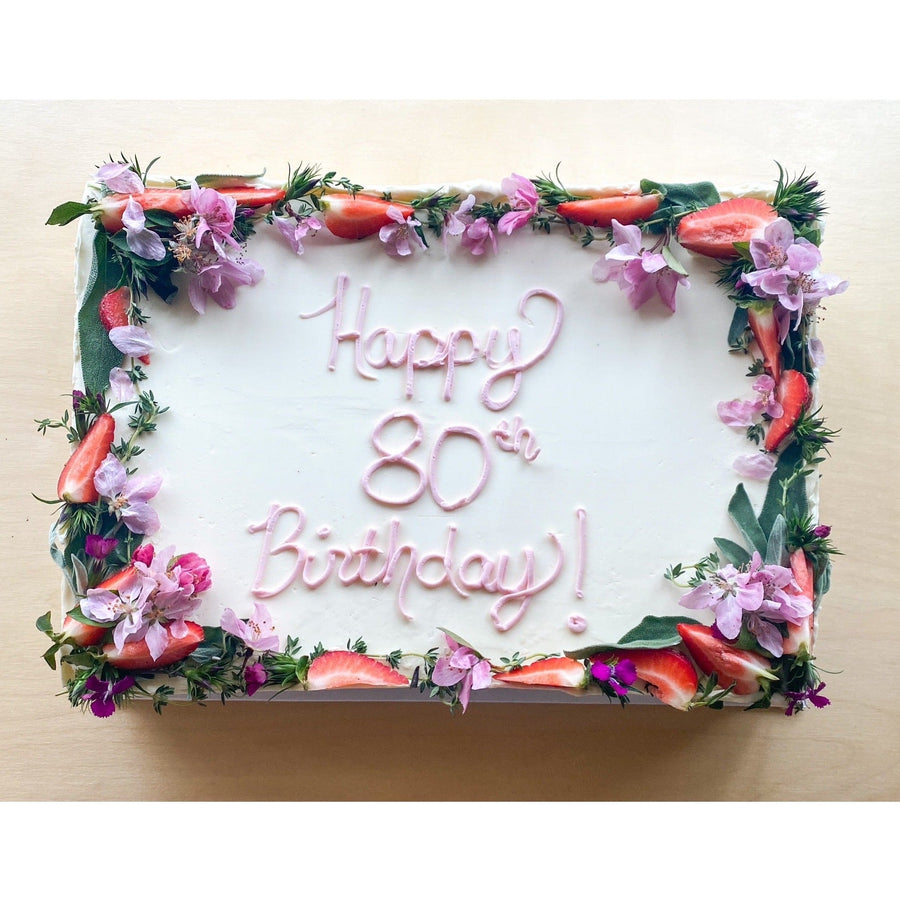 A white sheet cake with a crown of fresh flowers and strawberries; "happy 80th birthday!" is piped in pink buttercream.