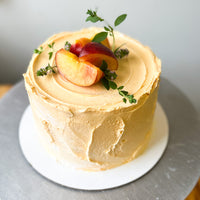 A round cake covered in light orange frosting and topped with fresh peach slices and basil sprigs.