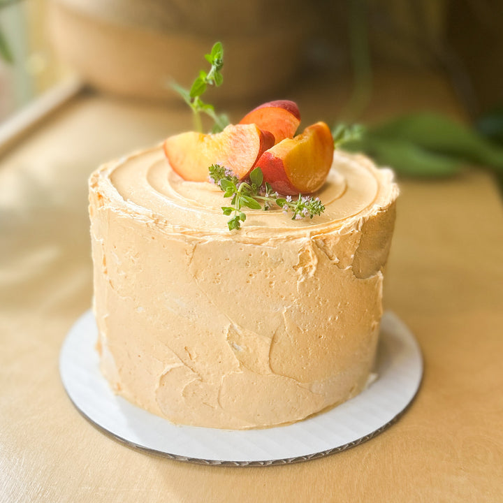 A round cake covered in light orange frosting and topped with fresh peach slices and basil sprigs.