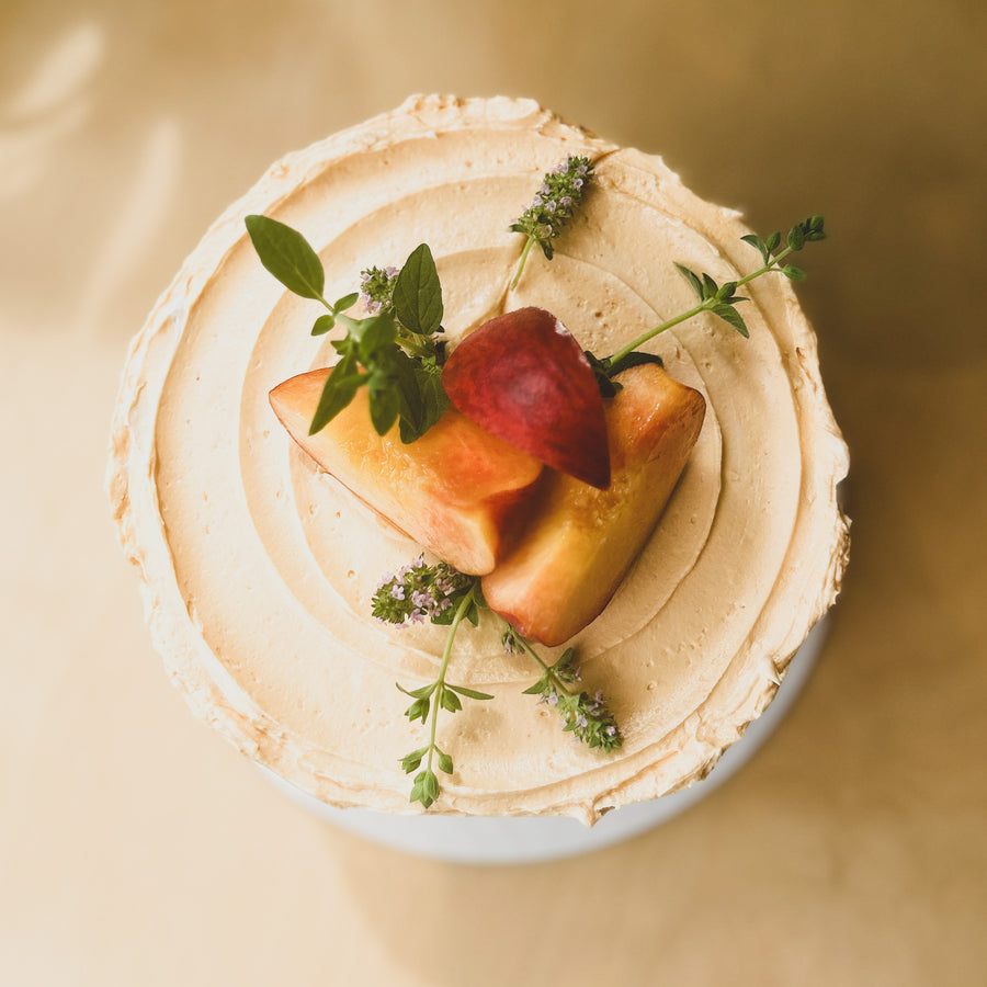 Top-down view of a round cake covered in light orange frosting and topped with fresh peach slices and basil sprigs.