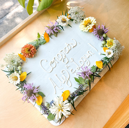 A sheet cake decorated along its border with fresh, pastel-toned flowers. Colors include white, yellow, and purple.