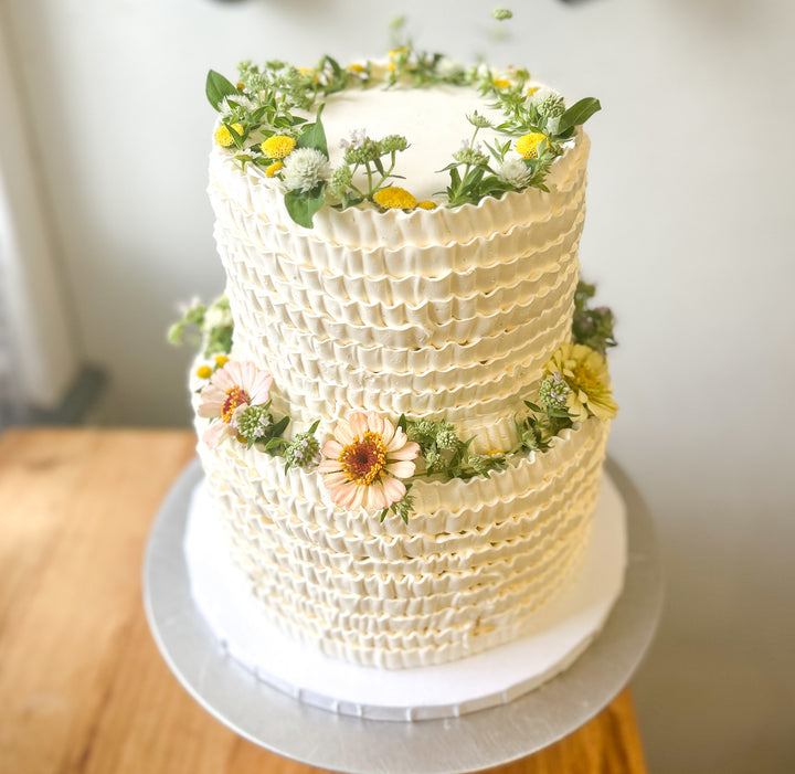 A two-tier cake decorated with crowns of fresh, pastel-toned flowers on each tier. Colors include light orange and yellow.