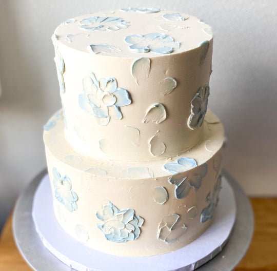 A two-tier cake frosted with white buttercream and decorated with blue, abstract buttercream flowers. 