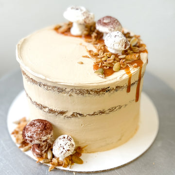 A round cake frosted with a sheer layer of light brown buttercream and decorated with a crescent of caramel drizzle, granola, and small mushroom-shaped meringues.