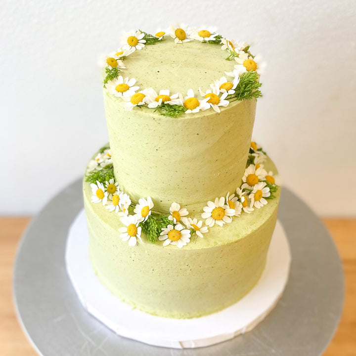 A two-tier cake frosted with light green buttercream and decorated with a fresh floral crown on each tier.