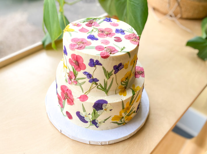 A two-tier cake decorated with brightly colored pressed flowers. Colors include pink, purple, and yellow.