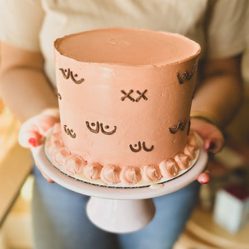 A cake frosted with light pink buttercream and decorated with cartoonish buttercream pairs of breasts, each unique. Some of the breasts have X's to represent mastectomies. 