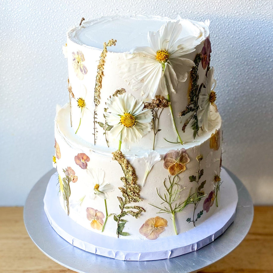 A two-tier cake frosted with rustic white buttercream and decorated with fresh and pressed florals.