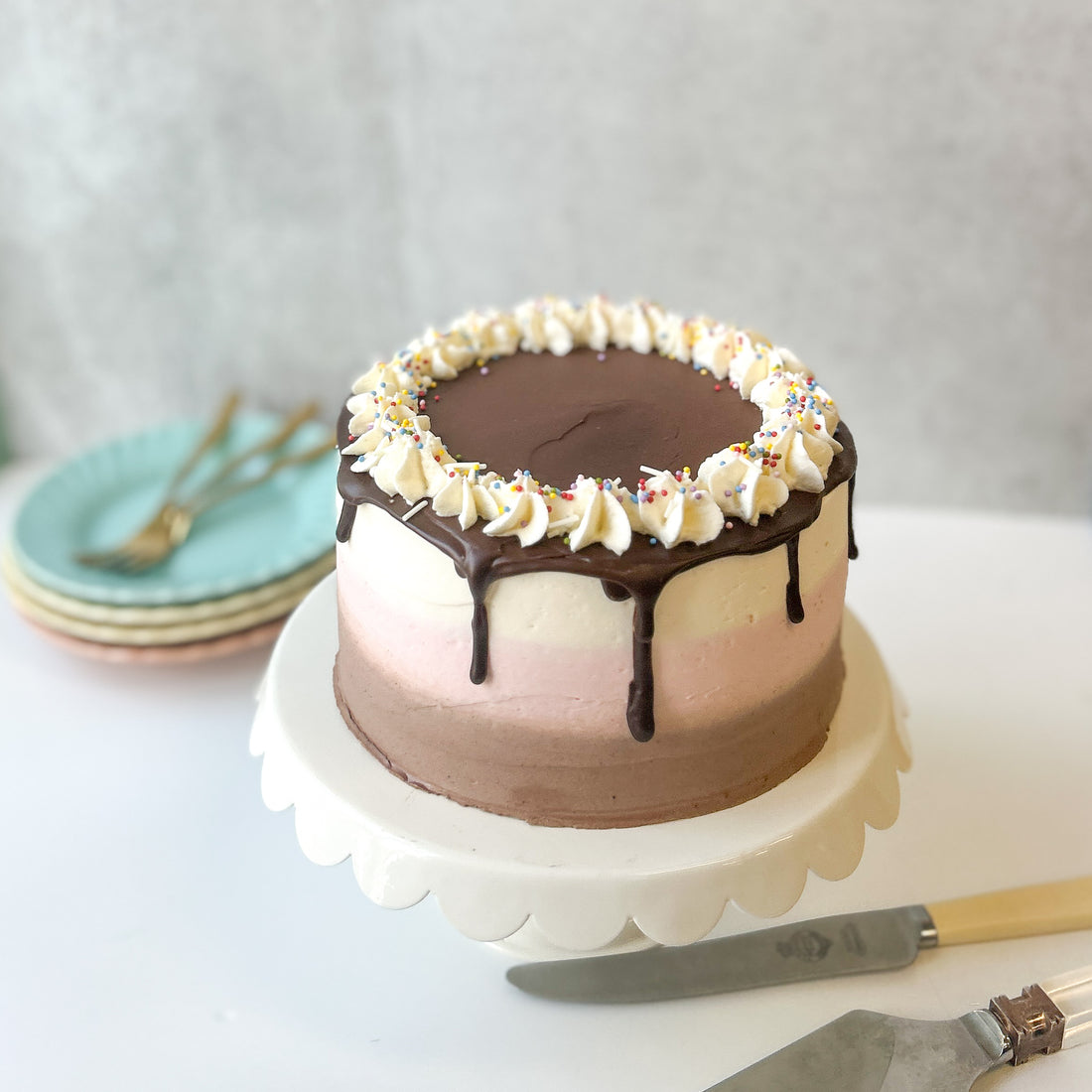 A round cake frosted with three bands of different flavors: vanilla, chocolate, and strawberry. The cake is topped with a chocolate ganache drip, a crown of whipped cream, and rainbow sprinkles.