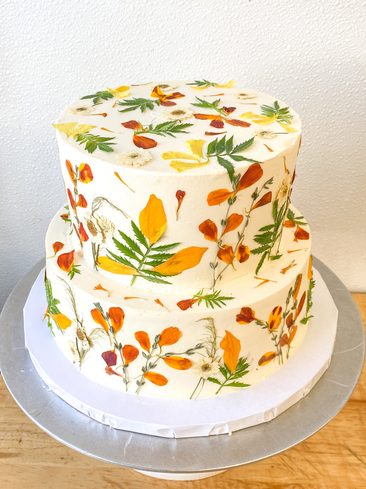 A two-tier cake decorated with brightly colored pressed flowers. Colors include orange, red, and yellow.