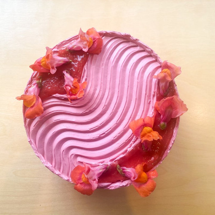 Top-down view of a cake frosted with hot pink buttercream. The frosting is piped in a wavy pattern across the top of the cake; beside the waves are pools of rhubarb compote and fresh snapdragon flowers.