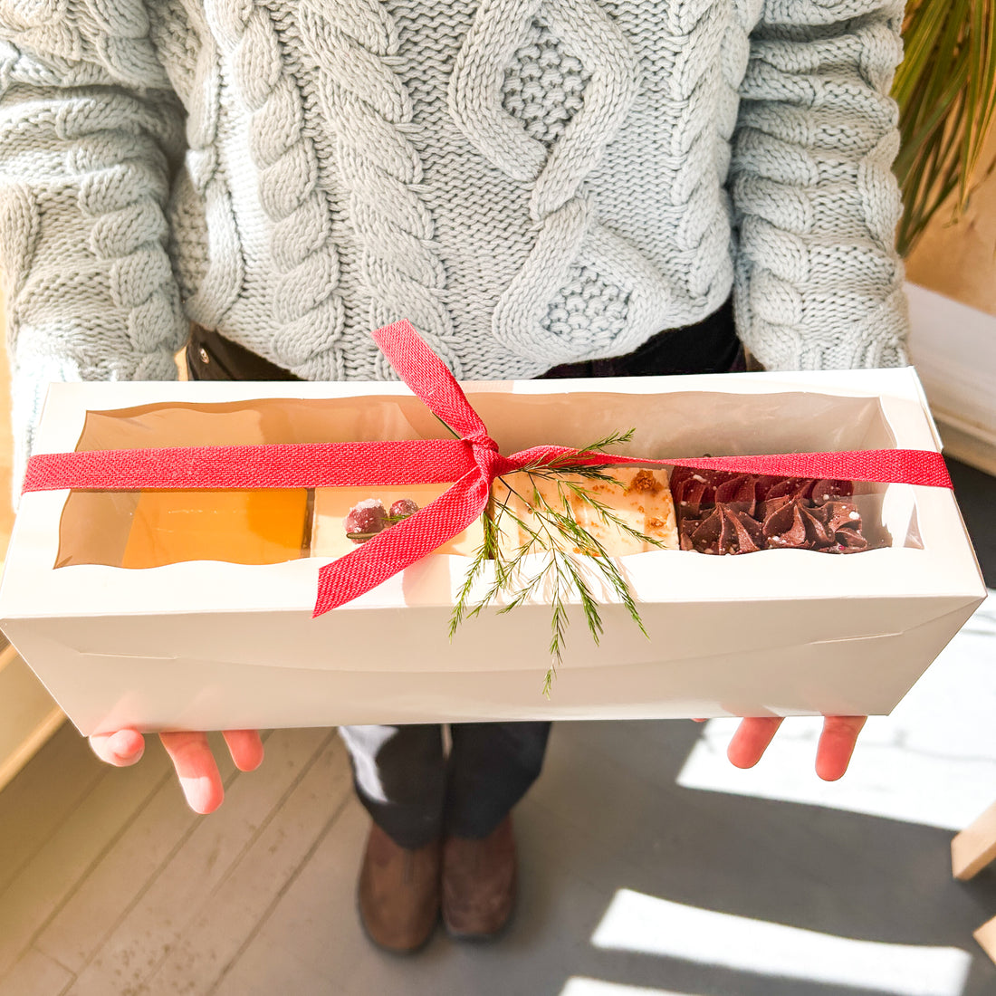 A person holding a white rectangular box tied with red ribbon; the box contains four square cakes, each a different flavor.