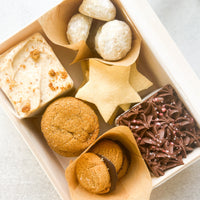 An open box containing a variety of sweets: two small square cakes (one chocolate, one vanilla), peanut butter cookies, shortbread stars, powdered sugar cookies, and ginger cookies.