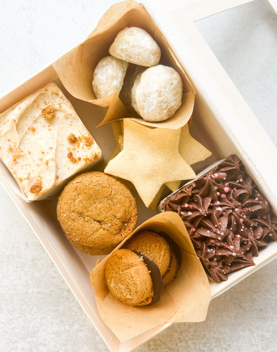 An open box containing a variety of sweets: two small square cakes (one chocolate, one vanilla), peanut butter cookies, shortbread stars, powdered sugar cookies, and ginger cookies.