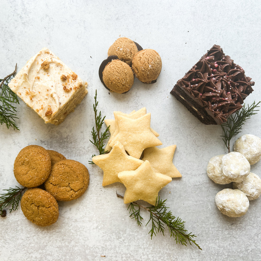 A variety of sweets laid out on a table. Clockwise from the center, they are: peanut butter cookies, a square chocolate cake, powdered sugar-coated "wedding" cookies, shortbread stars, ginger cookies, and a square vanilla cake.