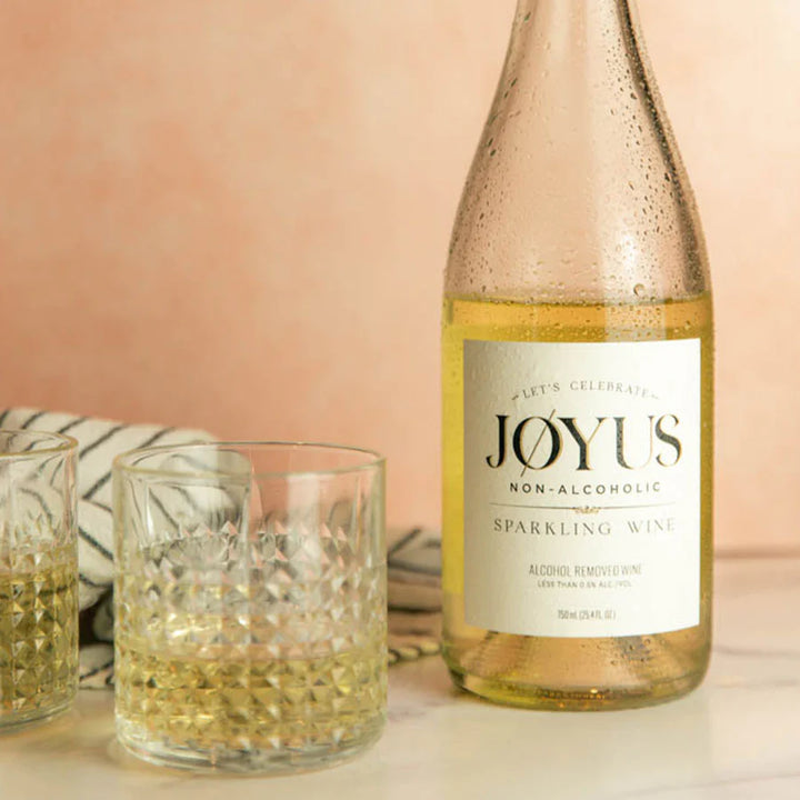 A bottle of white wine beside two glasses of the same. The label of the wine reads: "Let's Celebrate. Joyus Non-Alcoholic Sparkling Wine. Alcohol Removed Wine."