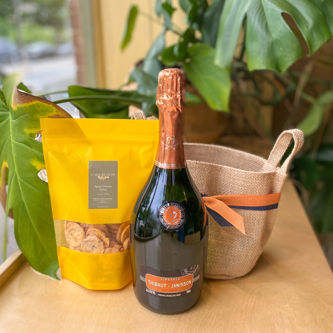 A bottle of Thibaut-Janisson, a bag of spicy cheese coins, and an empty tote bag tied with a blue and orange ribbon.