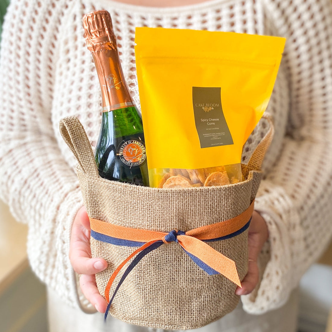 A person holding a small burlap tote containing a bottle of Thibaut-Janisson wine and a bag of spicy cheese coins. The tote is tied with orange and blue ribbon.