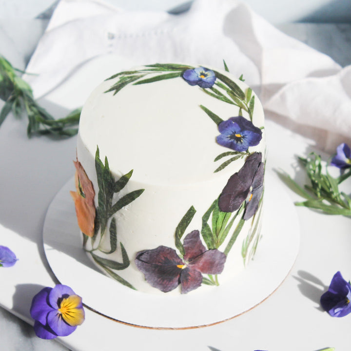 A cake decorated with smooth white buttercream and crescents of pressed flowers along the side and top.