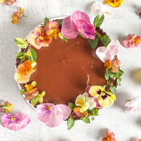 Top-down view of a cake covered in ganache and chocolate crumbs. The top of the cake is decorated with a crown of fresh flowers and greenery.