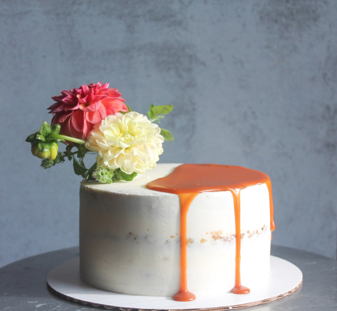 A round cake covered in a sheer layer of white buttercream; it is topped with a bouquet of flowers on one side and a partial pool of caramel on the other.