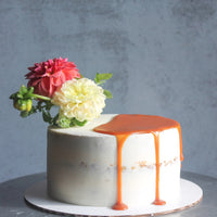 A round cake covered in a sheer layer of white buttercream; it is topped with a bouquet of flowers on one side and a partial pool of caramel on the other.