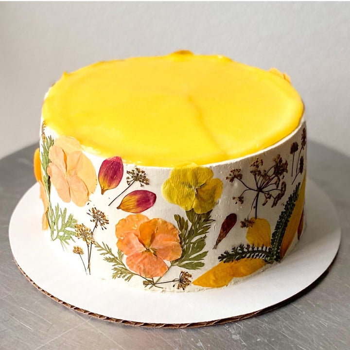 A round cake covered in white buttercream. The top of the cake is covered in lemon curd; the sides of the cake are decorated with pressed flowers.