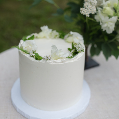 A tall round cake frosted with smooth white buttercream and decorated with a white floral crown.