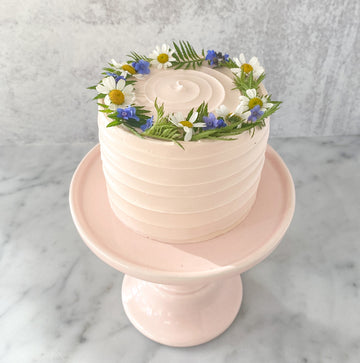 A round cake frosted in light pinked buttercream, which has been applied in a spiral pattern top to bottom. It is also decorated with a fresh flower crown.