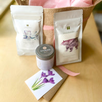 Two pouches of loose-leaf tea, a small notecard, a jar of sugar cubes, and a small burlap tote bag that is filled with tissue paper and tied with a pink ribbon.