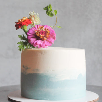 A cake frosted in white buttercream, with a diffuse band of light blue buttercream along the bottom. The cake is topped with a tall bouquet of flowers. 