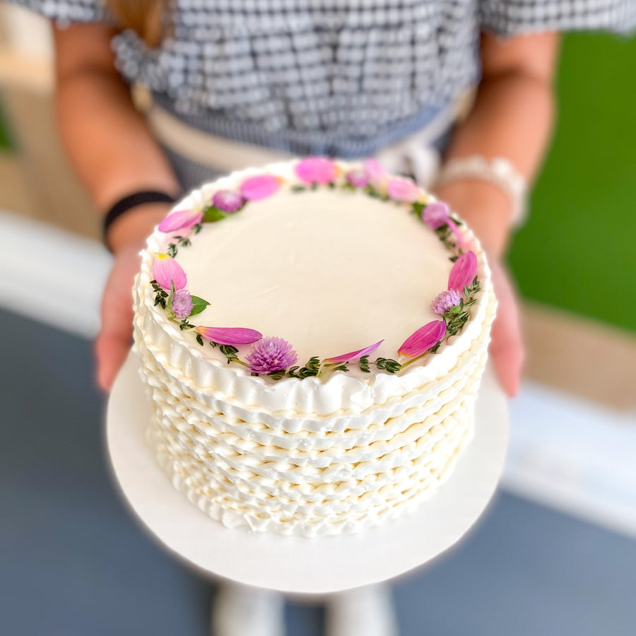 A round cake decorated with white buttercream ruffles along the sides and topped with a crown of flower petals.