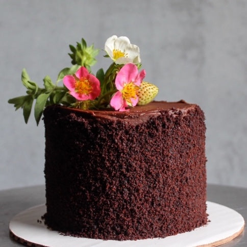 A round cake covered in ganache and coated in chocolate crumbs along its side. The top of the cake is decorated with a bouquet of fresh flowers and greenery.