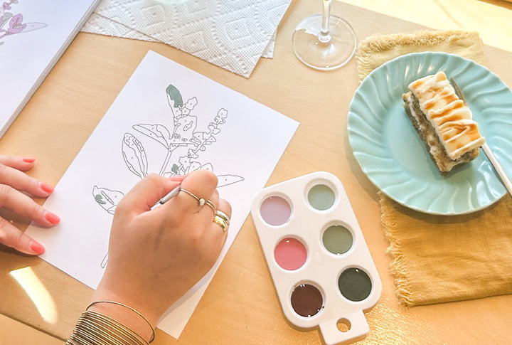 A woman painting a paint-by=numbers picture of flowers. There's a slice of cake on a plate beside her palette.