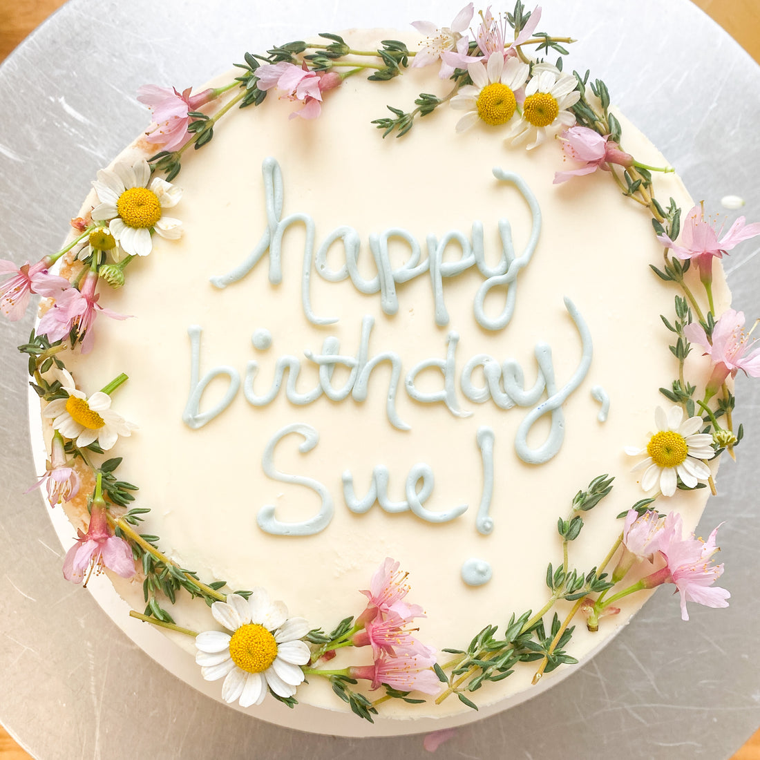 Top-down view of a round cake decorated with a flower crown and the words "happy birthday Sue!" in blue cursive piping.